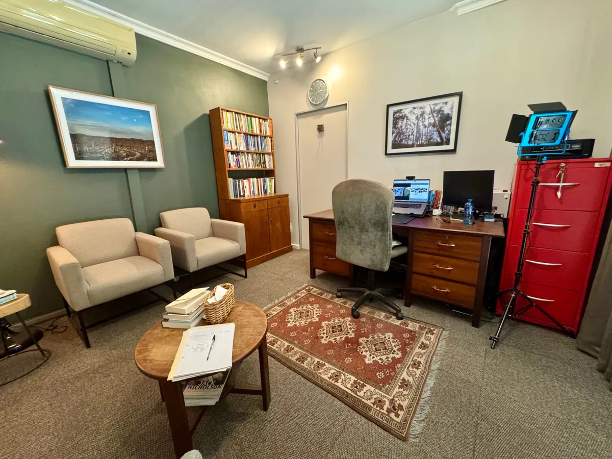 The counselling room from the other side: two cream armchairs facing a coffee table and a rug, with a bookshelf and a framed landscape