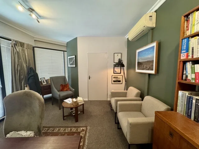 The counselling room from the desk: two armchairs along the green wall, a reading chair by the window and a small round table